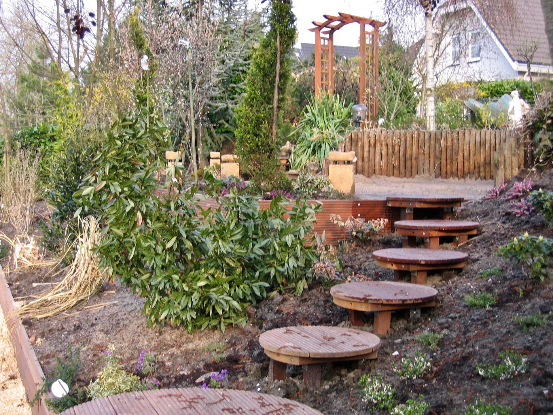 Tuin met houten trappen en platforms die een helling op leiden naar een pergola en een schutting.