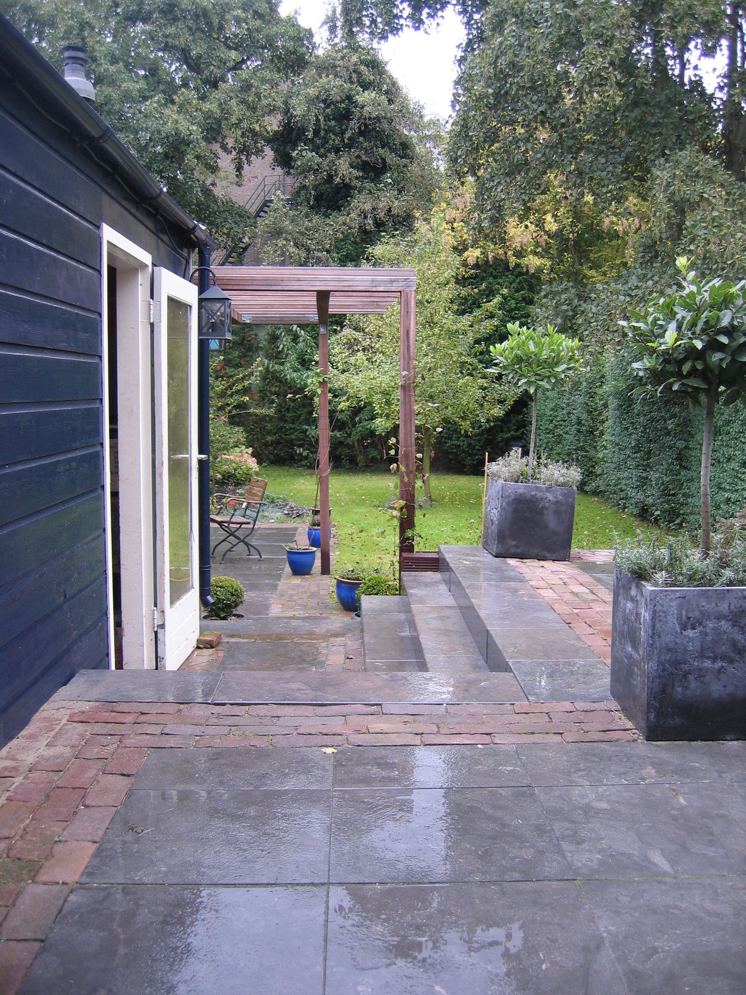 Terras in de achtertuin met een trap naar een grasveld, een houten pergola en een donkerblauwe schuur.