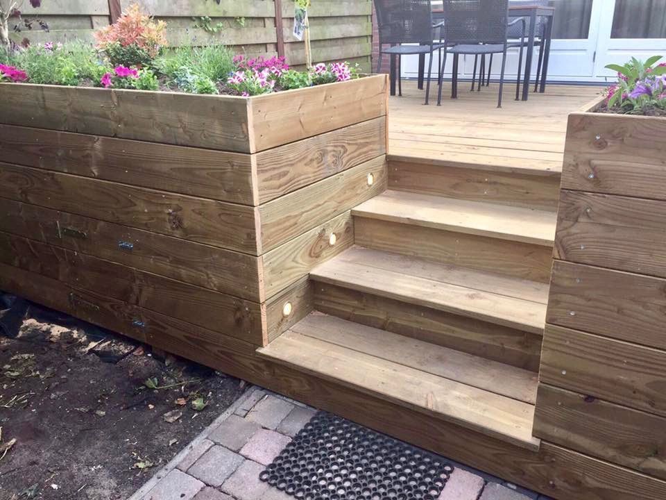 Houten plantenbak met trapje naar een terras. Bloemen in de plantenbak, lampen aan de muur.