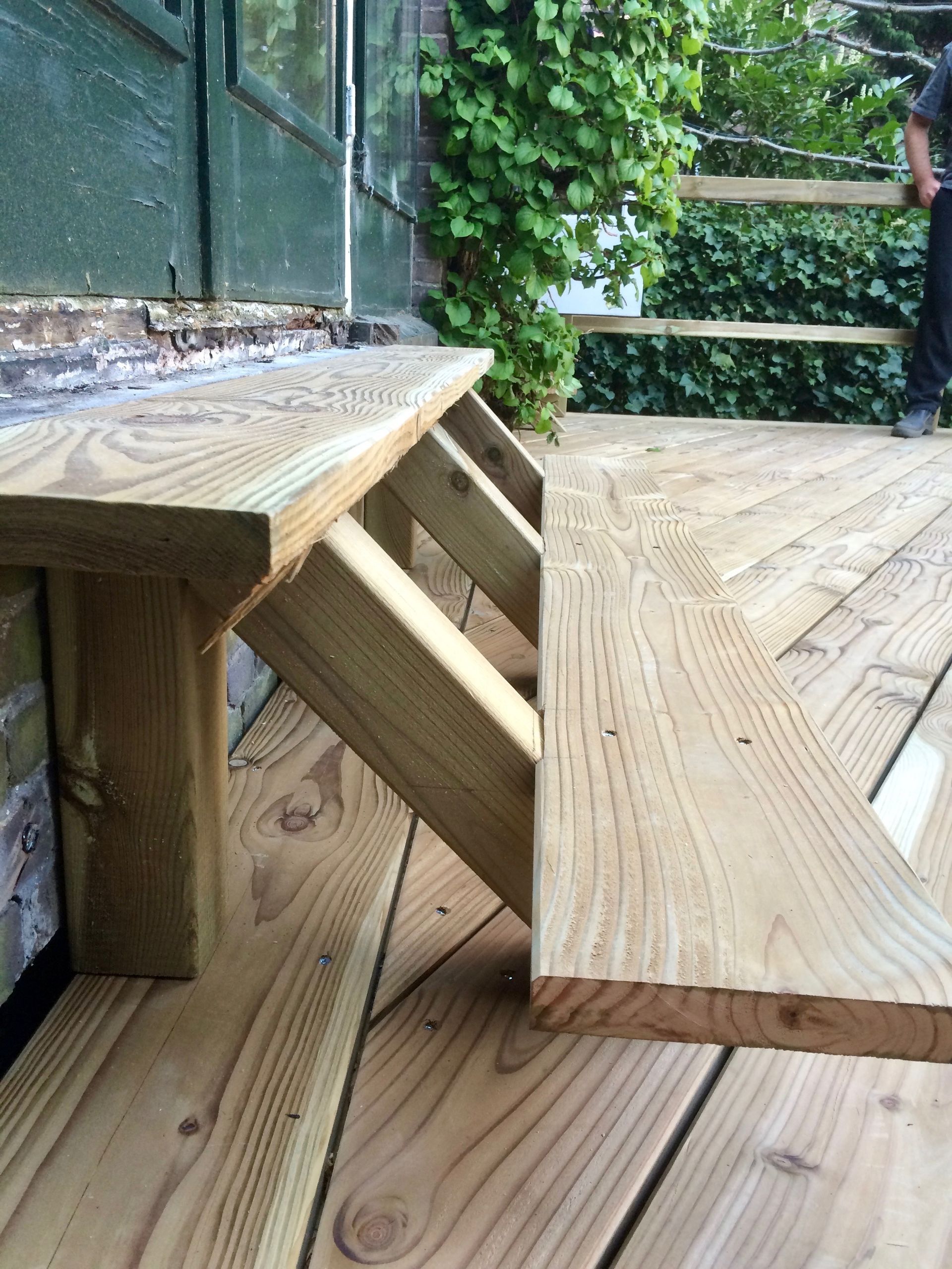 Houten bank op het terras. De terrasplanken zijn licht van kleur. Een persoon staat er vlakbij. Groen gebladerte op de achtergrond.