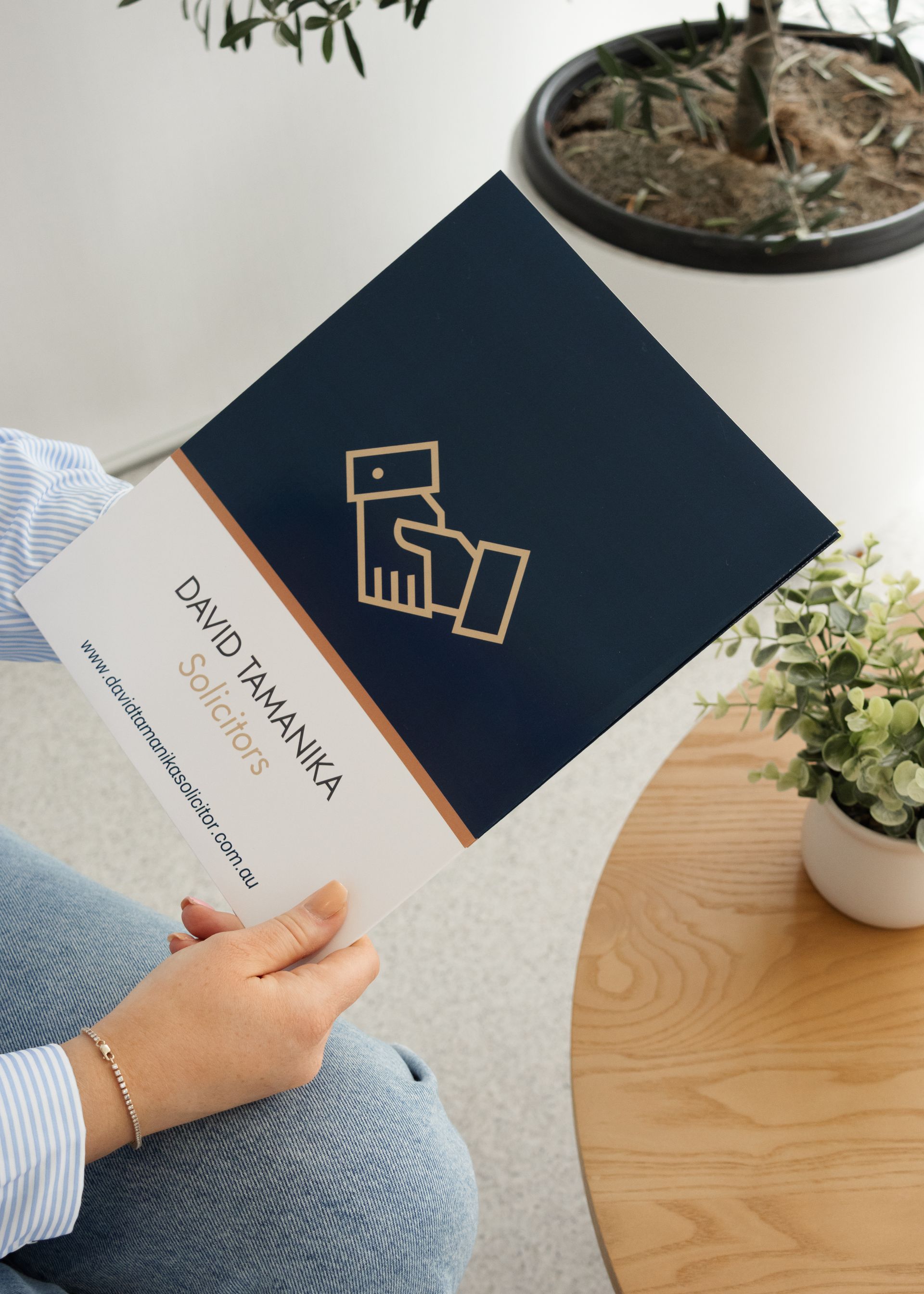 Person Holding a Navy and White Brochure at A Wooden Table with A Plant — David Tamanika Solicitors in Ballarat Central, VIC