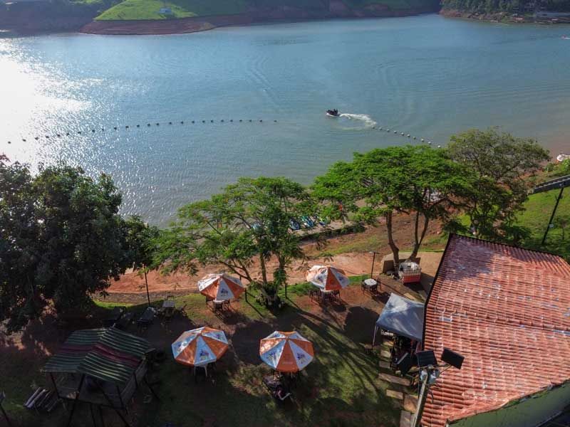 Cena à beira do lago: jet ski na água, guarda-sóis de praia, árvores e telhado de telha vermelha. Dia ensolarado.