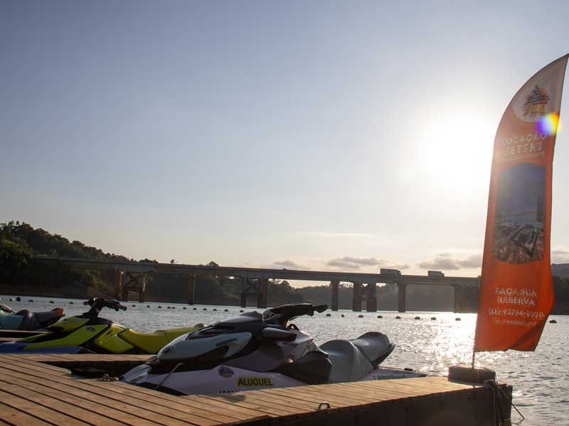 Jet skis atracados em um píer com uma ponte ao fundo sob um sol brilhante.