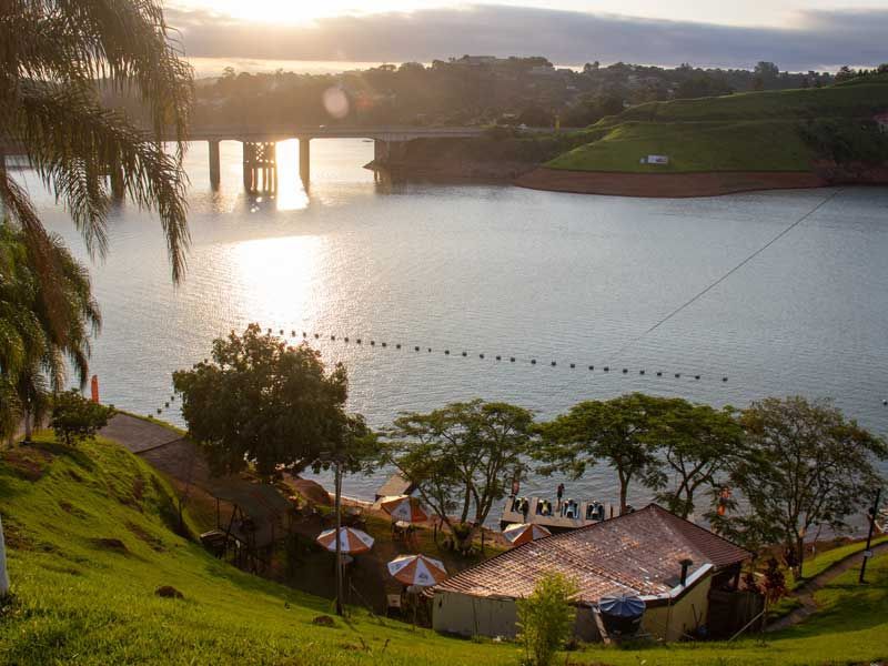 Pôr do sol sobre um lago com uma ponte, colinas gramadas e um pequeno prédio com guarda-sóis.