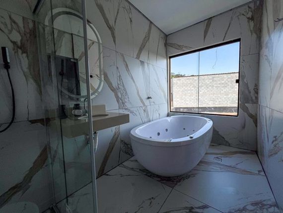 Banheiro moderno com banheira de hidromassagem branca, pia com bancada e paredes revestidas com azulejos de mármore. Uma janela oferece vista para o exterior.