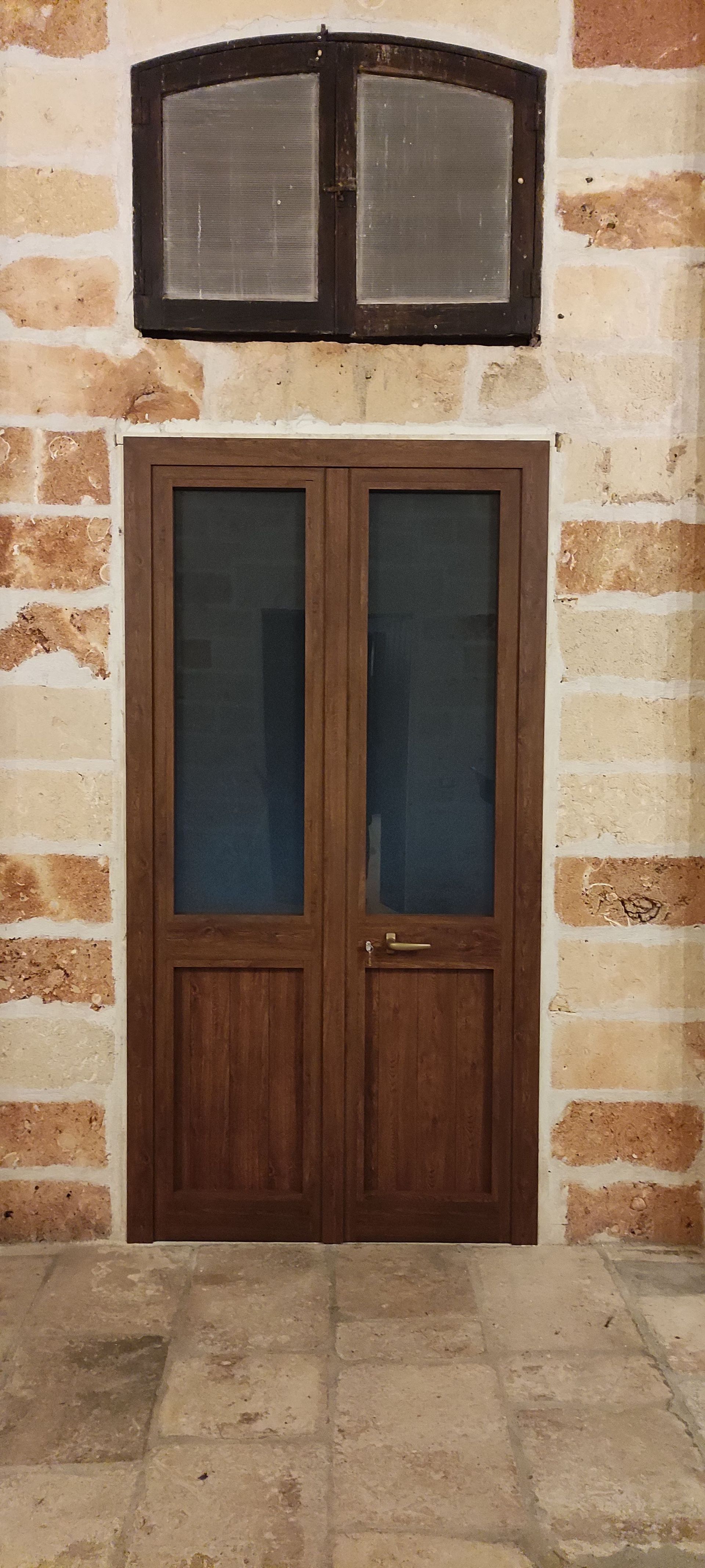 Una doppia porta in legno con pannelli in vetro è incastonata sotto una finestra a lunetta ad arco in una parete di pietra chiara e ruvida.