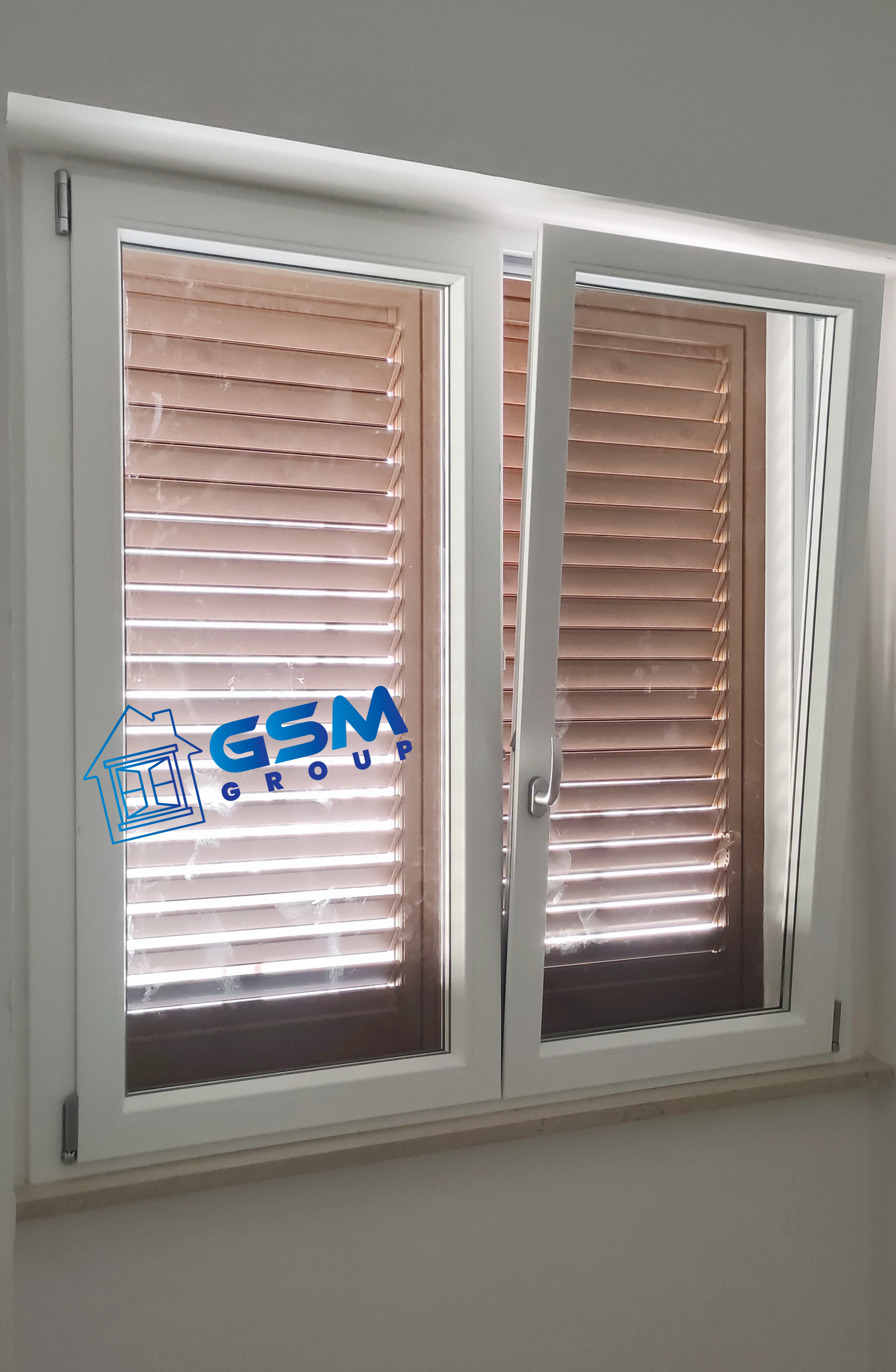 Finestra bianca a doppio vetro con persiane in legno all'interno, parzialmente aperta, con il logo blu del Gruppo GSM sul vetro.