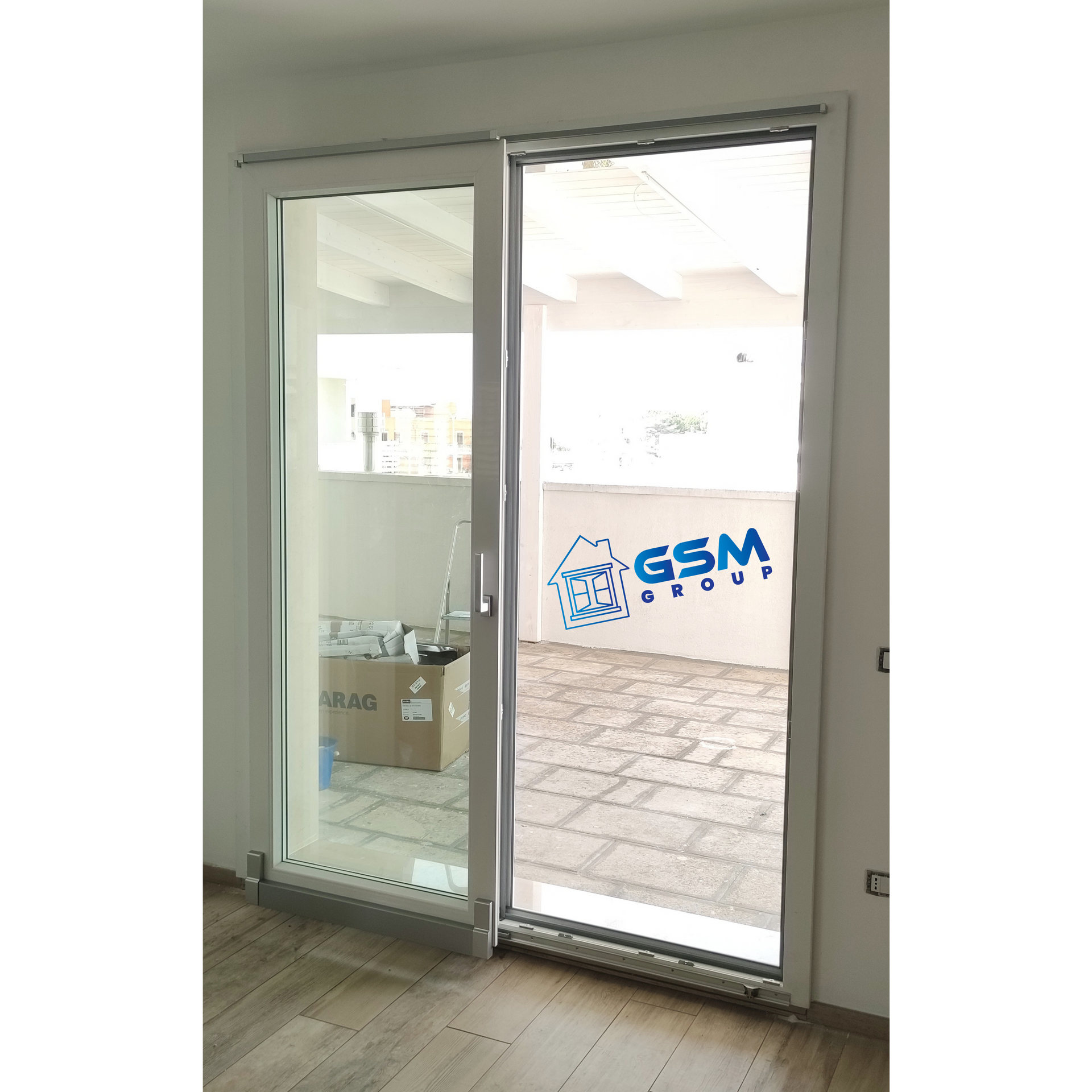 Una porta finestra con cornice bianca si apre su un patio esterno piastrellato in pietra, all'interno del quale è visibile una scatola di cartone.