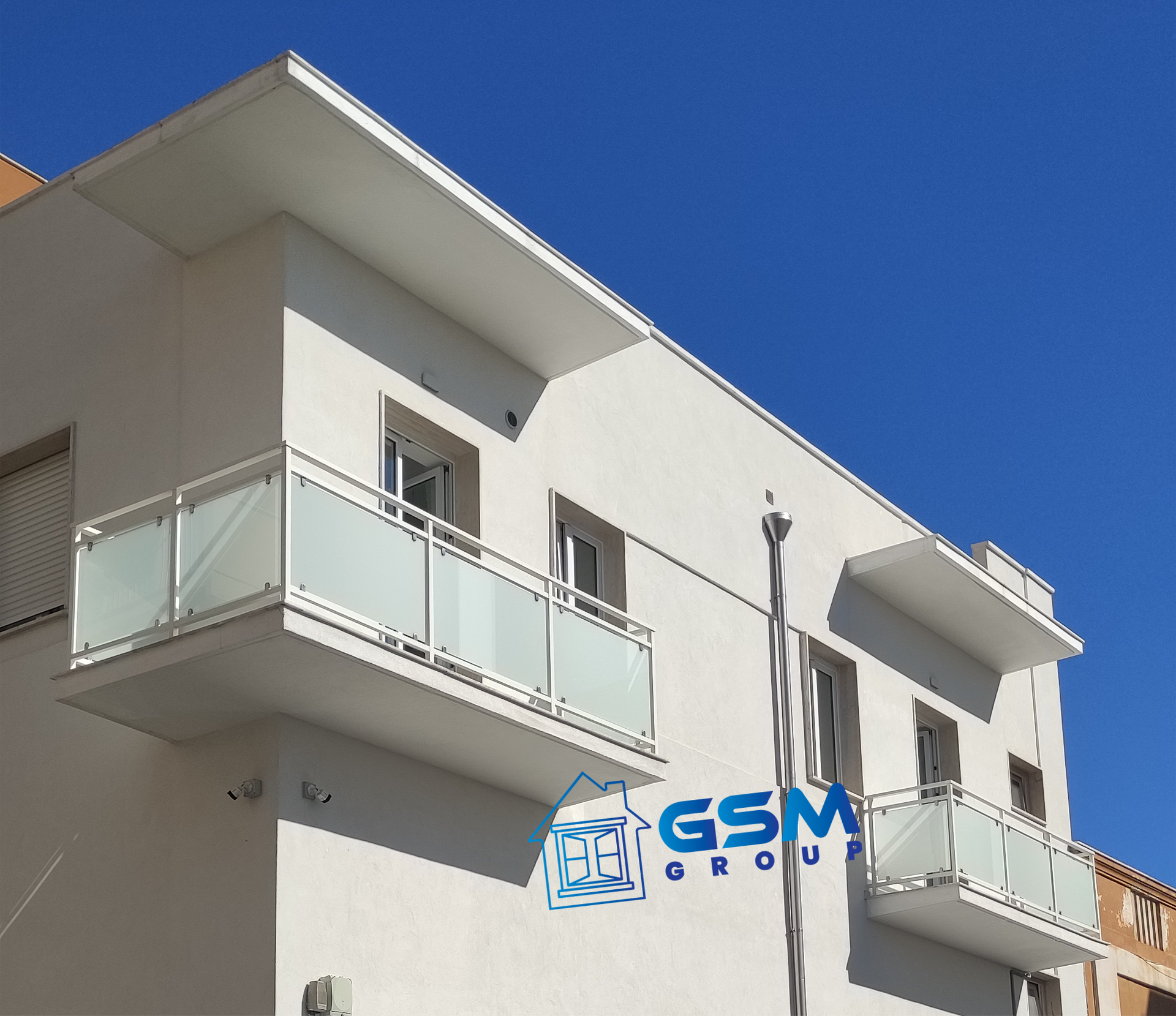 Un edificio bianco con moderni balconi a vetrata sotto un cielo azzurro limpido, su cui campeggia il logo del Gruppo GSM.