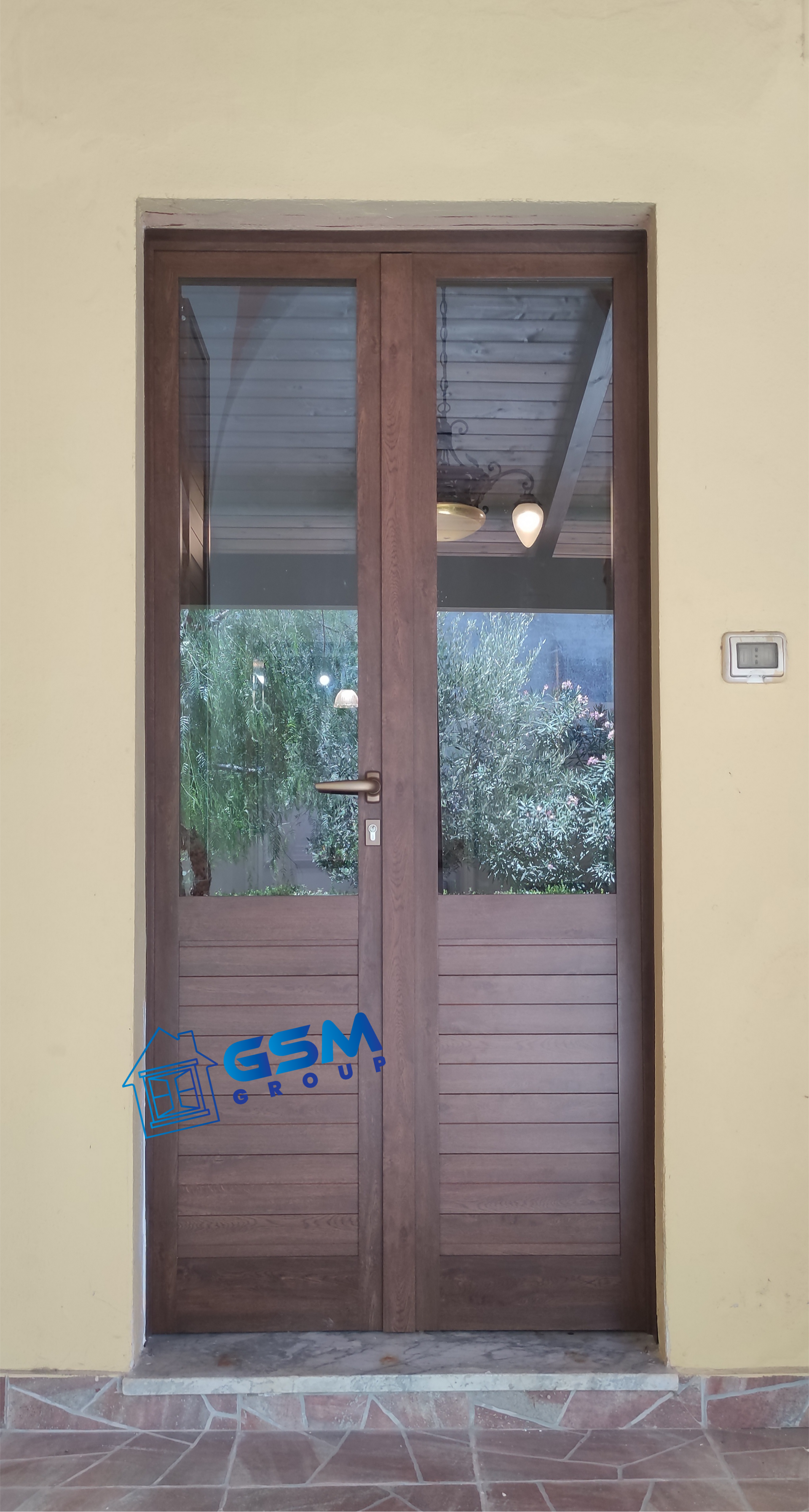 Una porta a doppio pannello marrone con sezioni superiori in vetro, incastonata in una parete beige, con una piccola tastiera digitale sulla destra.