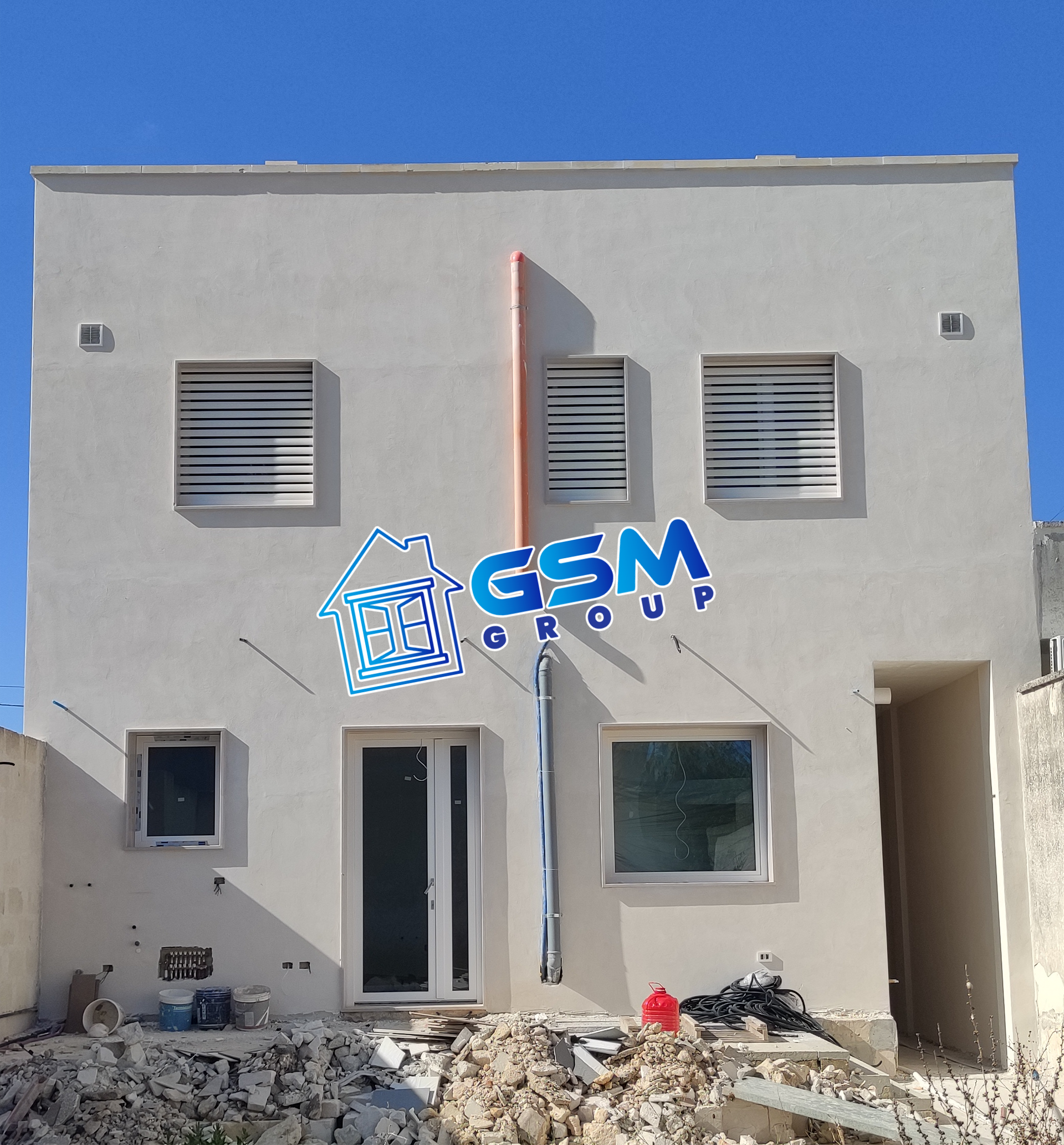 Un edificio residenziale a due piani, di colore beige, attualmente in costruzione, con persiane bianche e il logo del Gruppo GSM al centro.