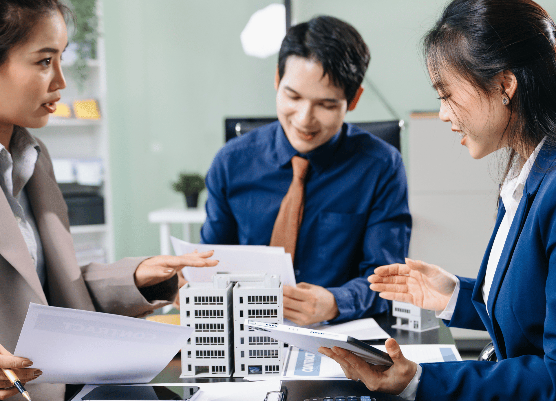 Three professionals in business attire collaborate at an office desk, discussing a white architectural building model.