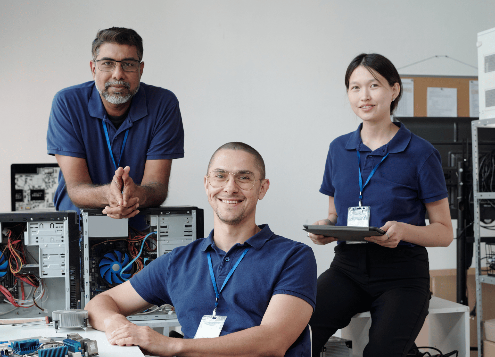 Three technicians in blue uniforms work in an IT lab with open computer towers, one holding a tablet.