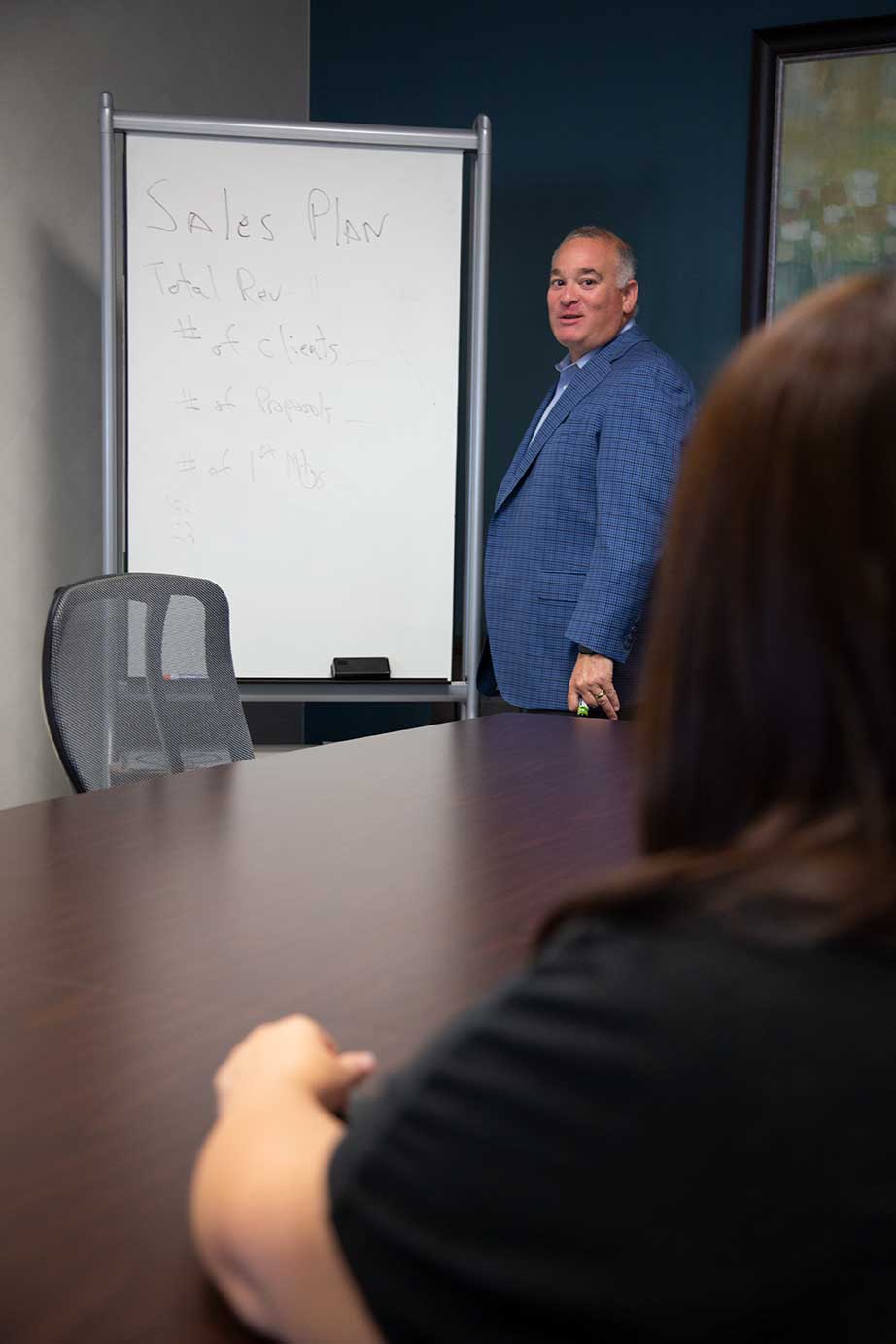 A person in a blue patterned blazer stands by a whiteboard in an office, facing someone across a conference table.