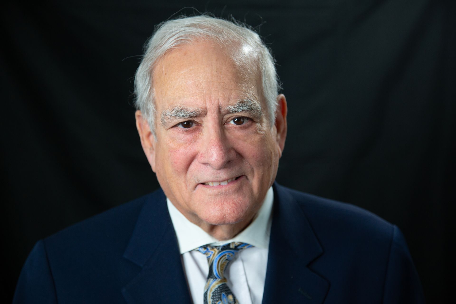 A headshot of a person wearing a navy blue blazer, white collared shirt, and patterned tie against a black background.