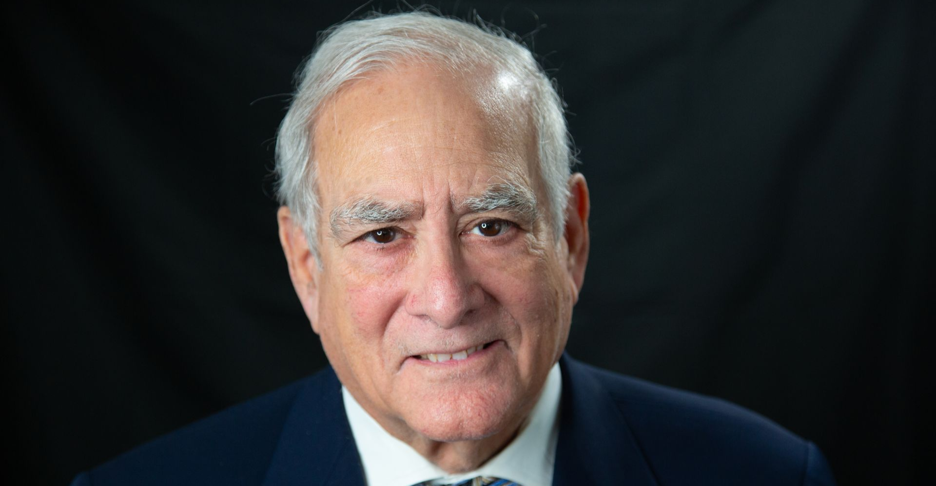 Headshot of a person with gray hair wearing a dark suit and white shirt against a black background.