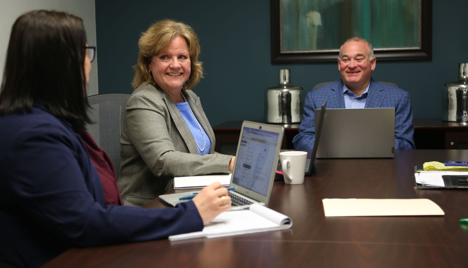 Three people sit around a boardroom table with laptops and documents, smiling during a business meeting.