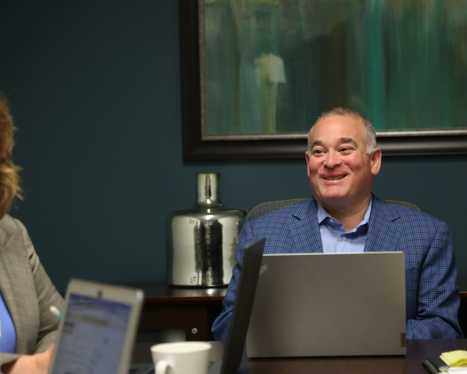 Man smiling behind a laptop in a conference room, with a coffee cup and blurred colleagues nearby