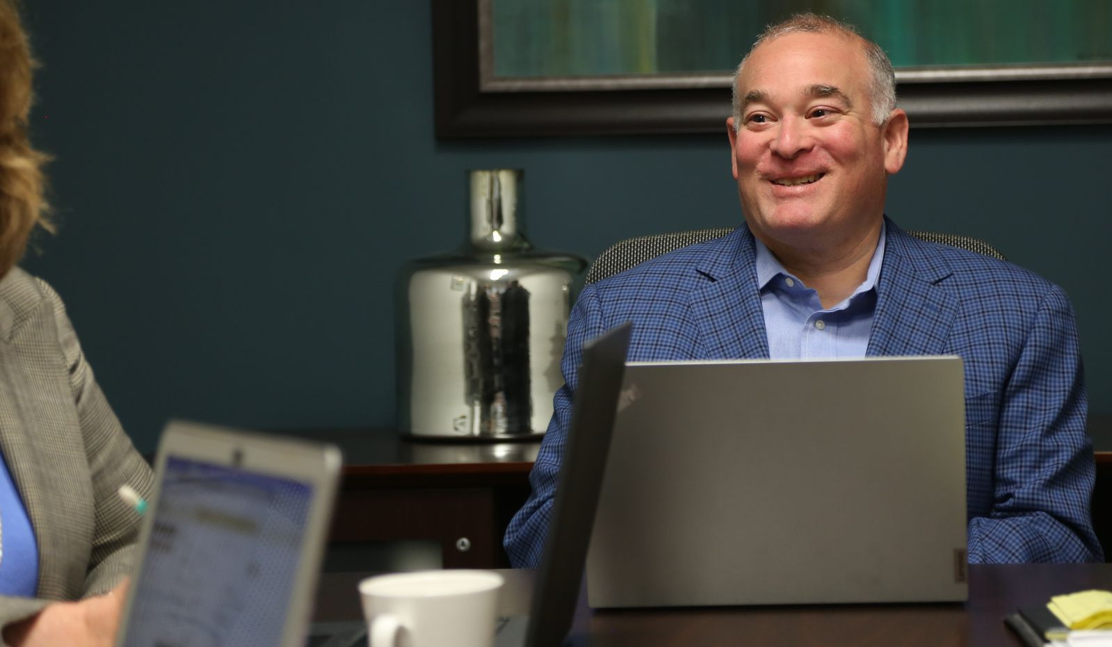 Smiling man in a blue blazer sits behind a laptop at a conference table, with a woman and coffee cup nearby