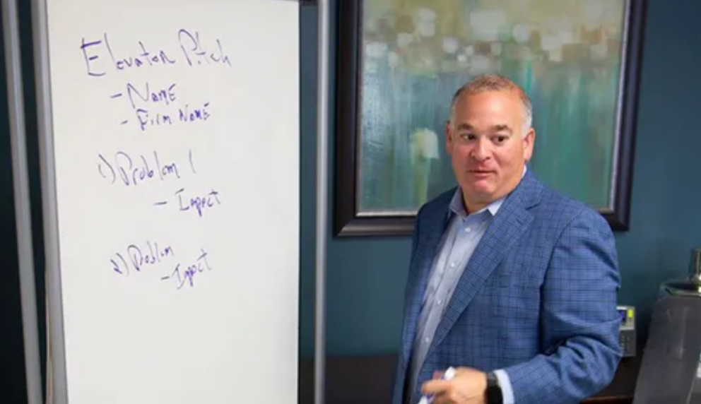 A person in a blue blazer stands by a whiteboard, gesturing toward an outline for an
