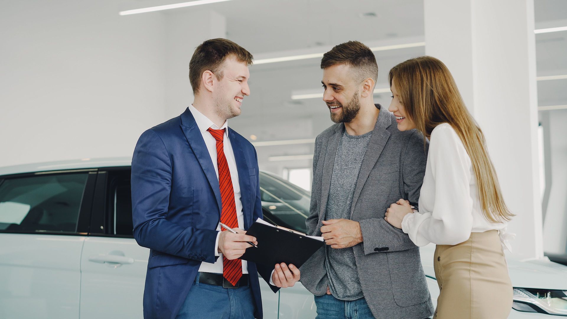 Salesman shaking hands with a couple in a car showroom, smiling beside a white SUV.