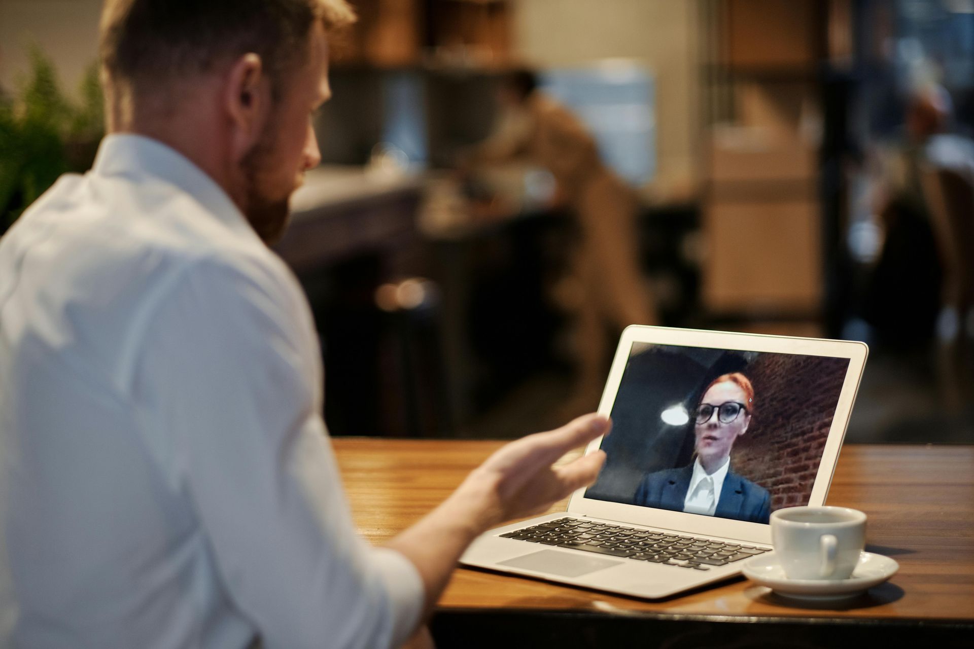 A person in a white shirt sits at a wooden table, gesturing during a video call on a laptop displaying a business meeting.