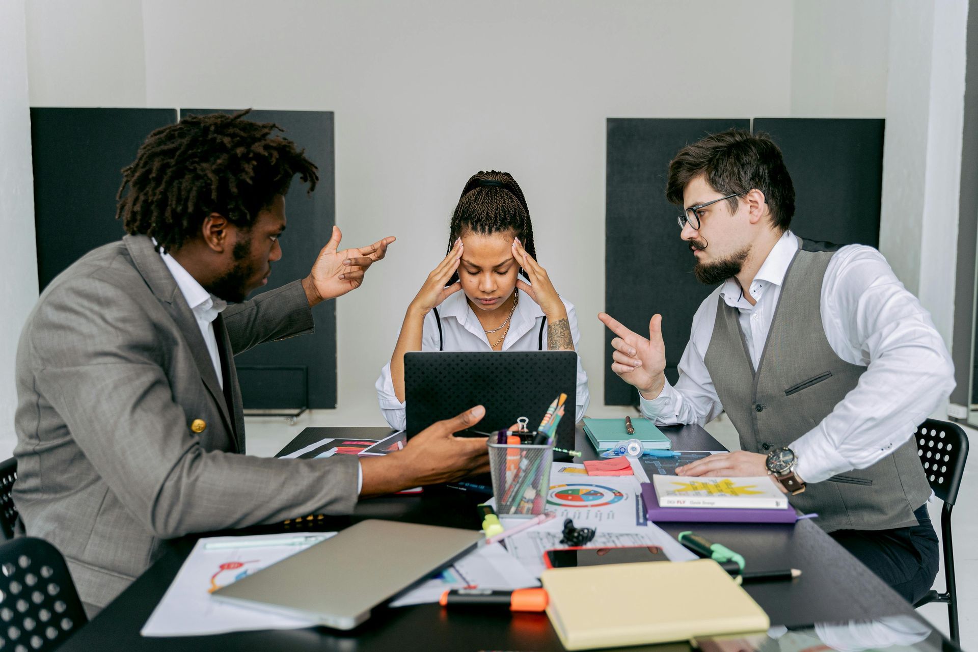 Two business professionals argue at a table while a colleague sits between them, holding their head in frustration.