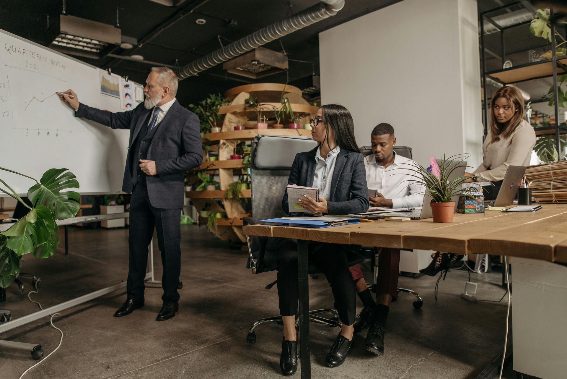 A professional leading a team meeting in a modern office, standing by a whiteboard while colleagues work at a wooden table.