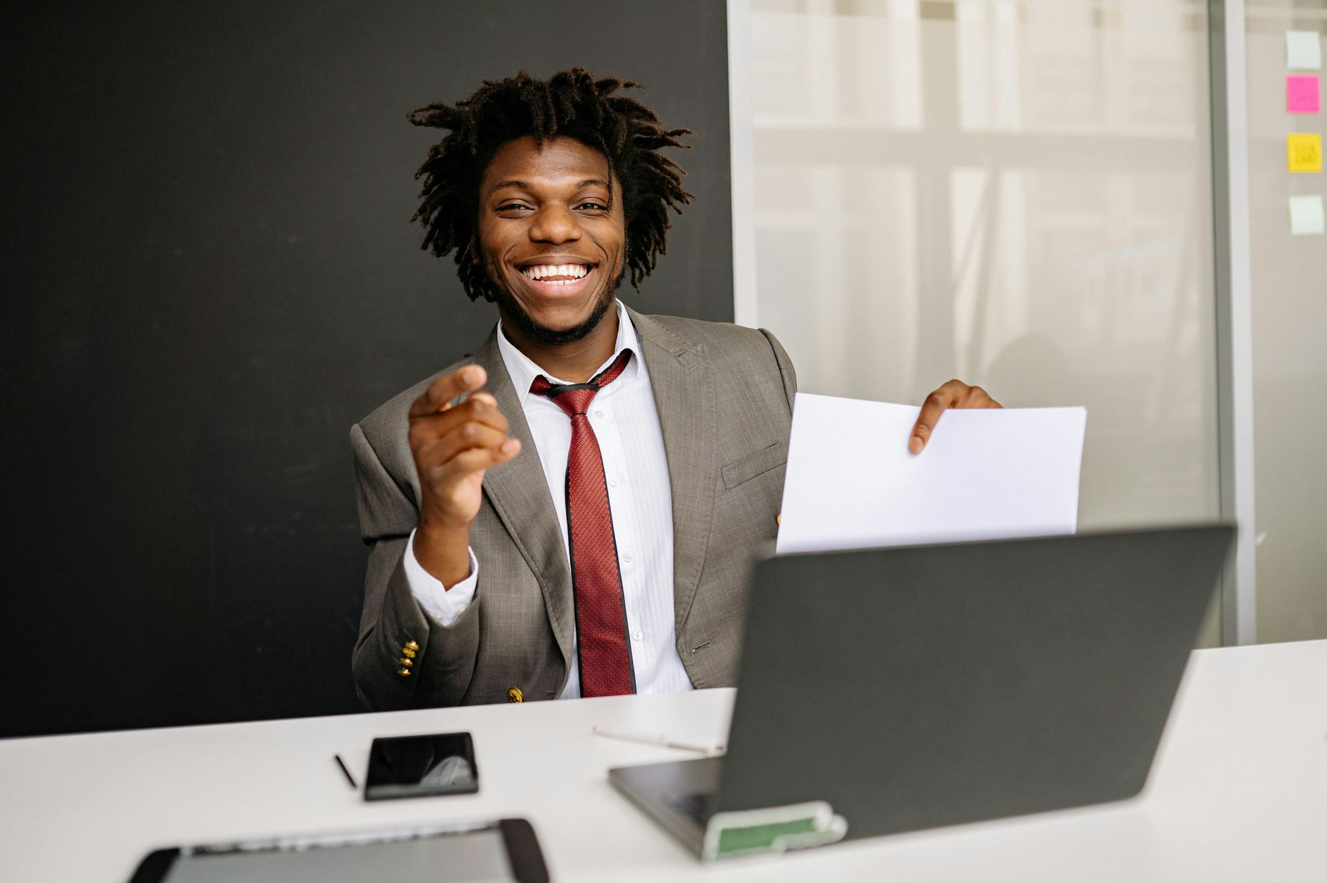 A smiling person in a suit and red tie sits at a desk, pointing forward while holding a document near a laptop.