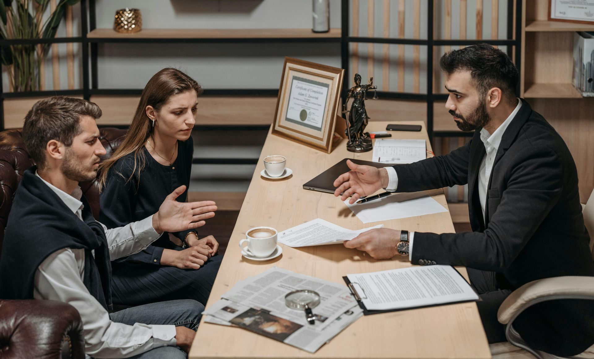 A professional meeting where a person gestures while explaining documents to two people across a desk in an office.