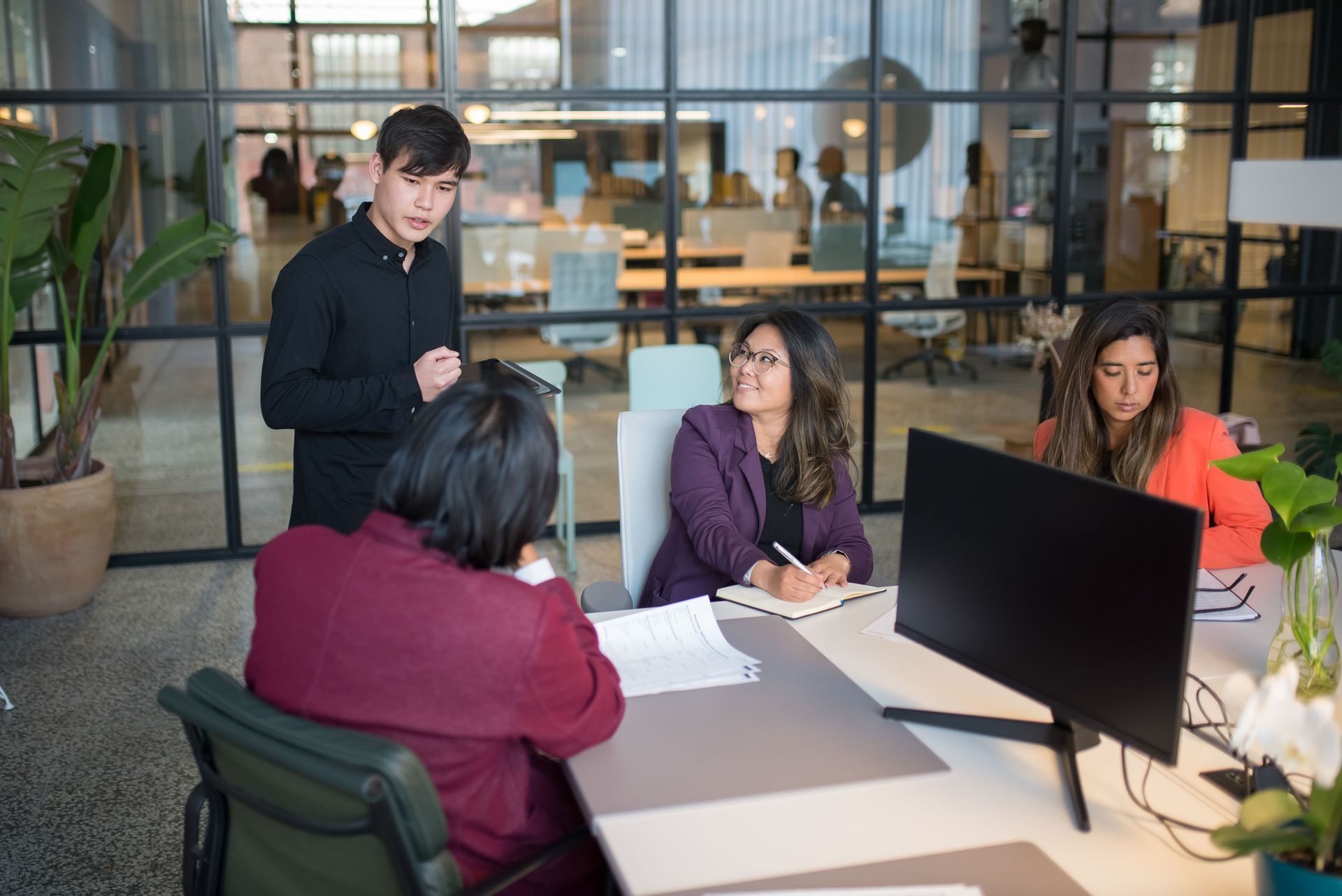 People meeting around a table in a glass-walled office, discussing papers beside a computer monitor.