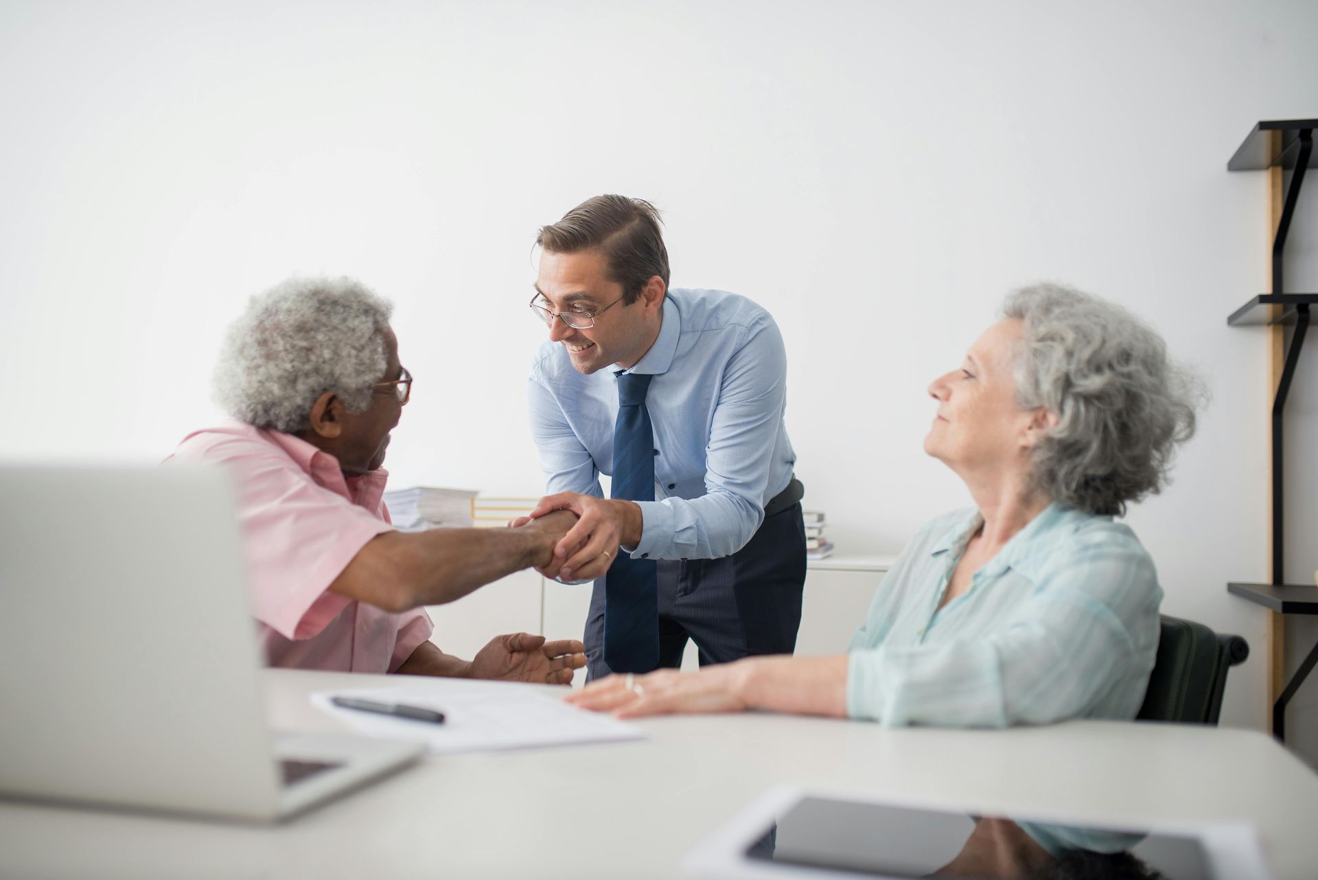 A professional shaking hands with a client while a second client looks on at an office desk with laptops.