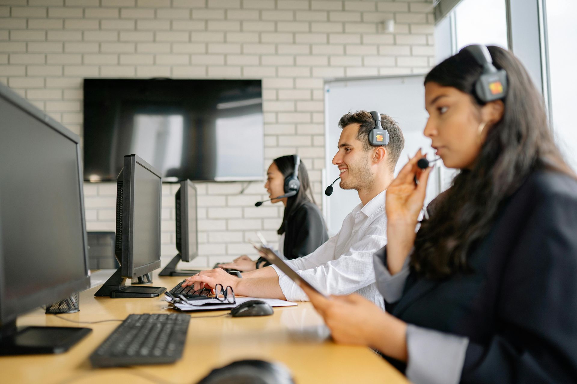 Three customer service representatives with headsets work at a desk in an office, focusing on their computer tasks.