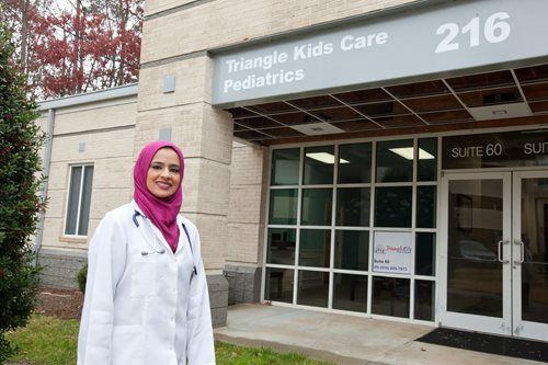 Woman in a white coat and hijab stands in front of a pediatric clinic entrance.