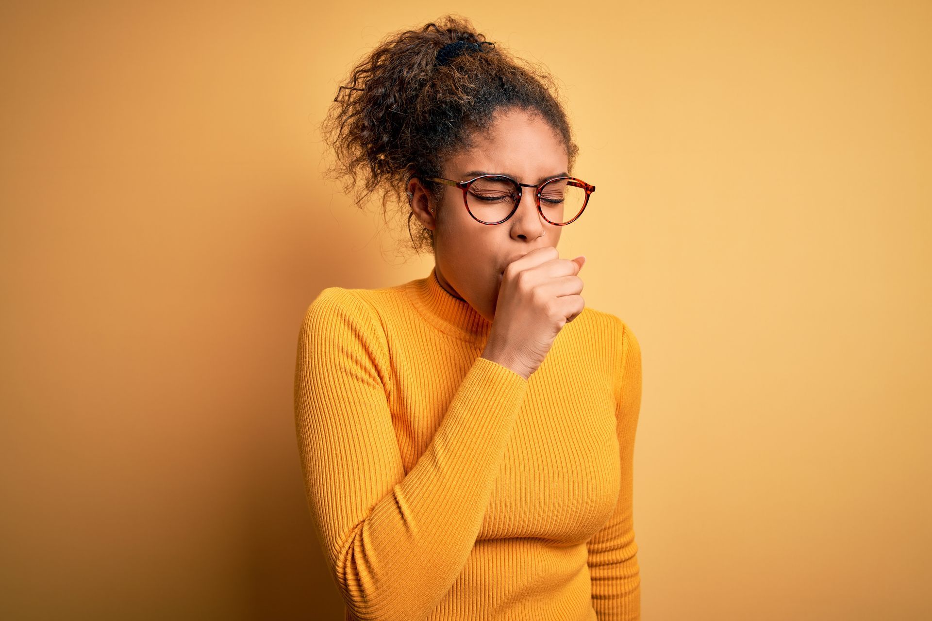 Woman coughing, covering mouth with hand, wearing glasses and yellow sweater, against a yellow background.