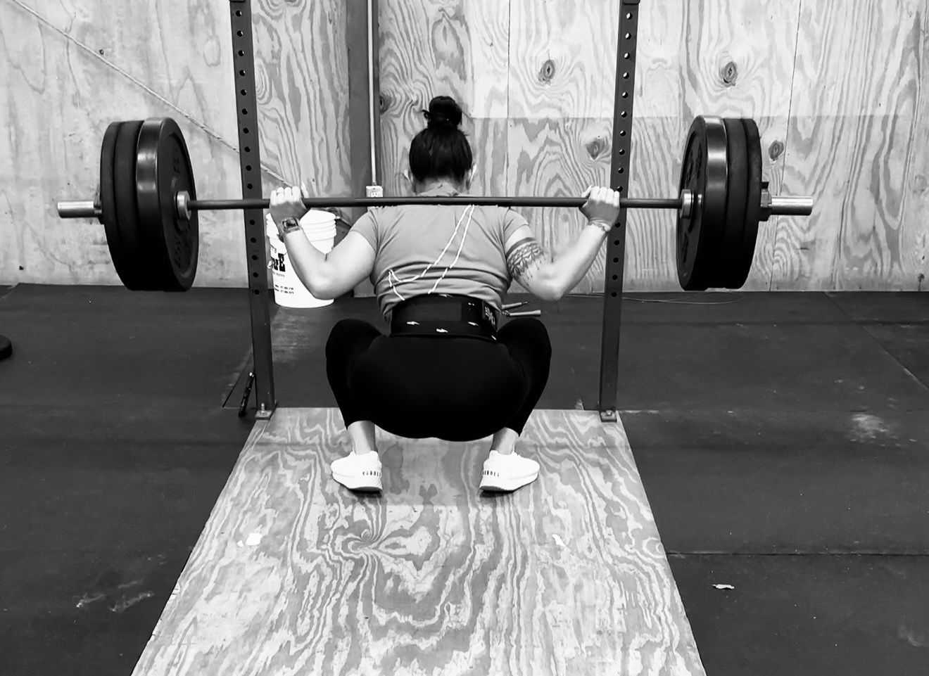 Woman performing a deep squat in a gym, back to the camera, lifting a barbell with weights. She wears a weight belt and is on a wooden platform.