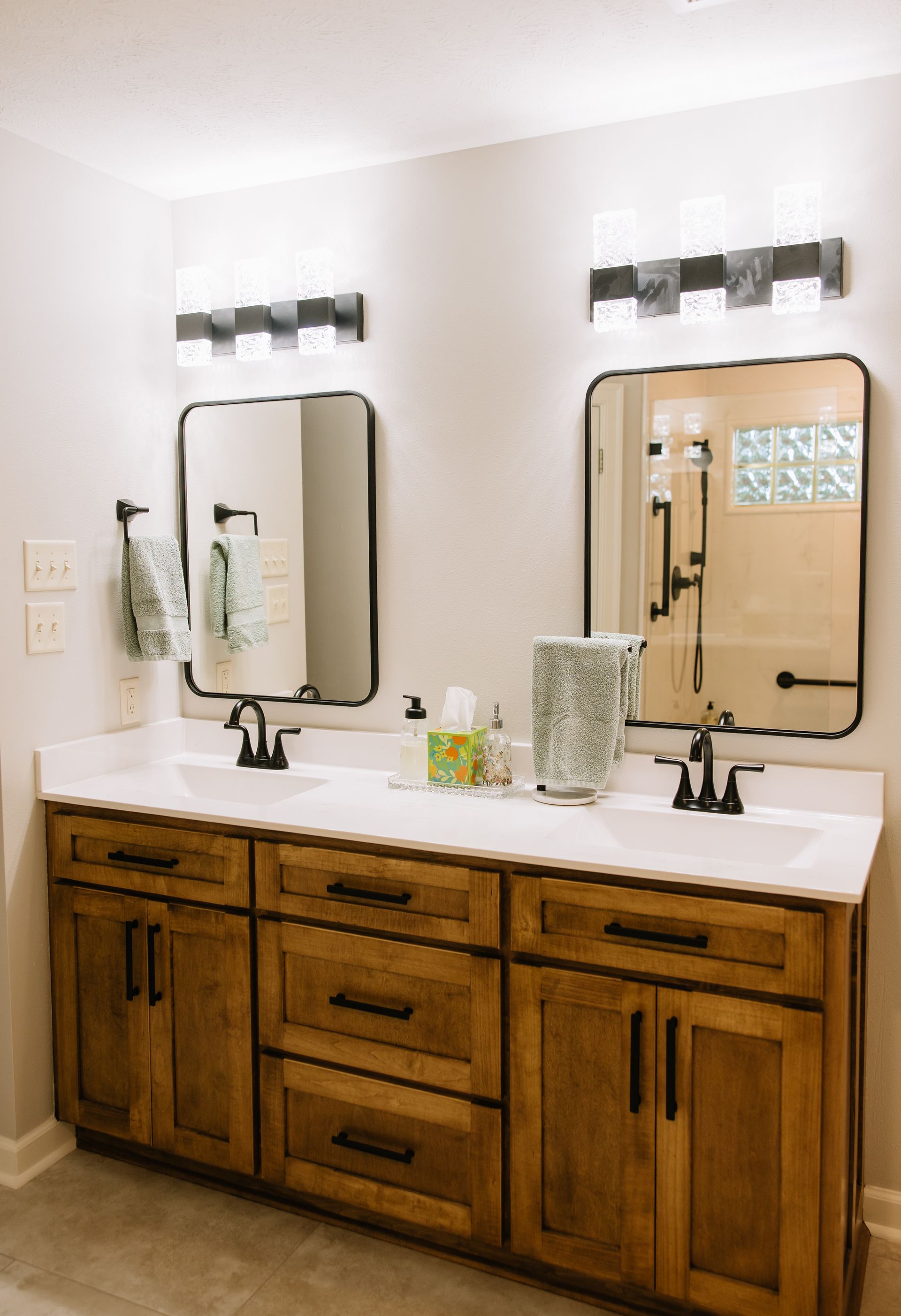 Bathroom vanity with double mirrors, white countertop, and wooden cabinets under wall sconces.