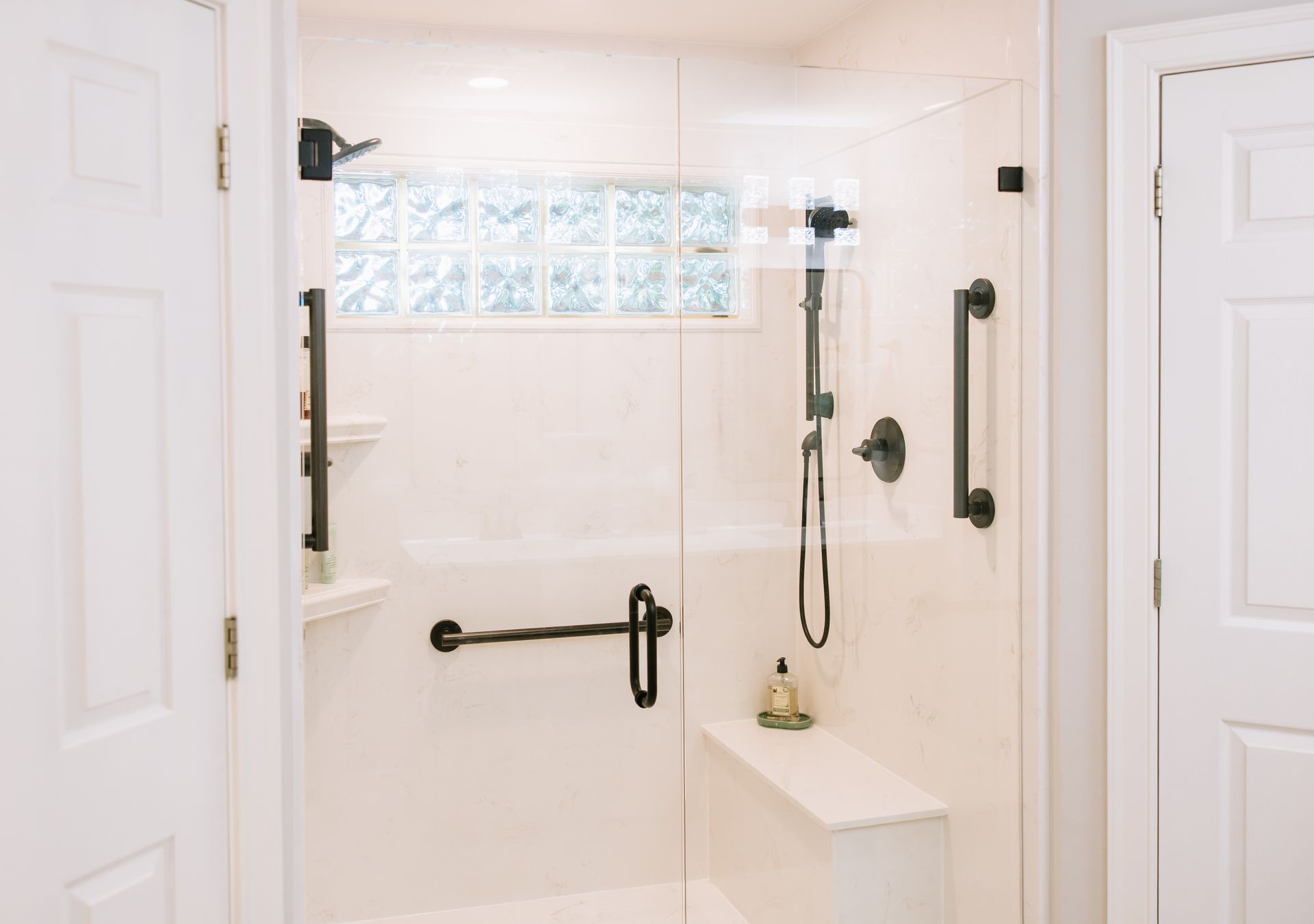 White accessible shower with grab bars, bench, and handheld showerhead in a bright bathroom.