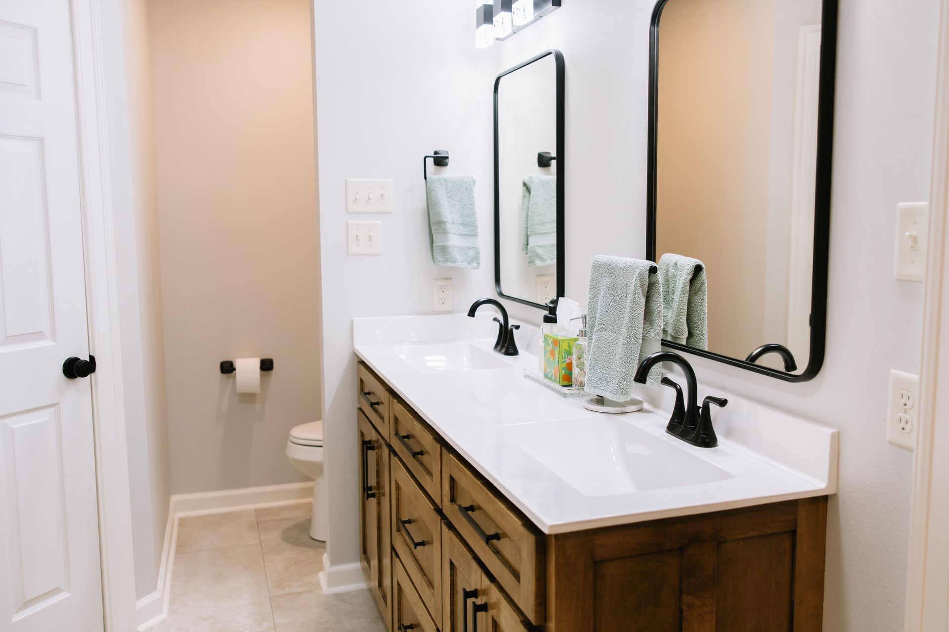 Bright bathroom with double vanity, mirrors, white countertops, and wood cabinets beside a toilet.