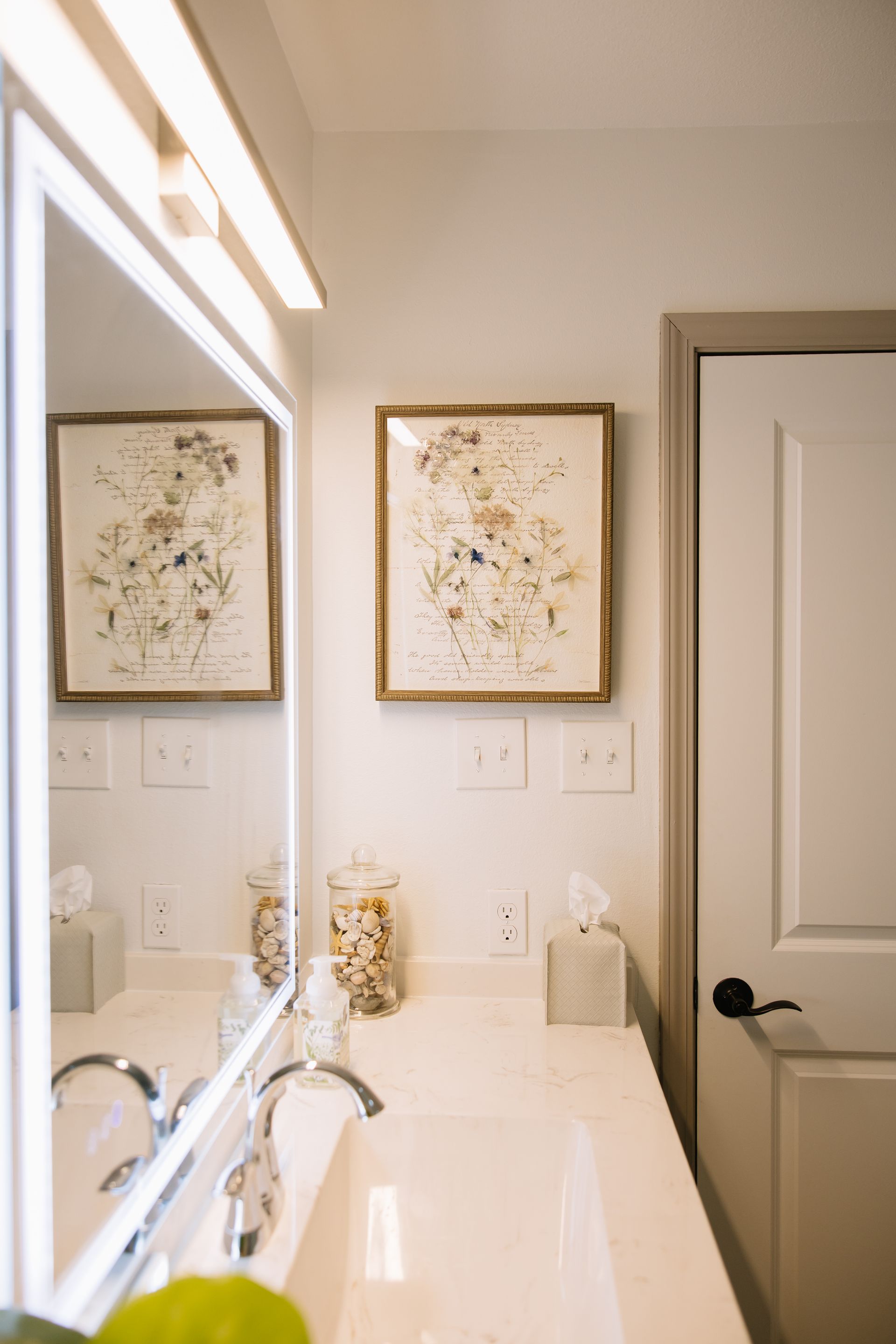 Bathroom vanity with large mirror, white countertop, framed floral art, and a closed door on the right.