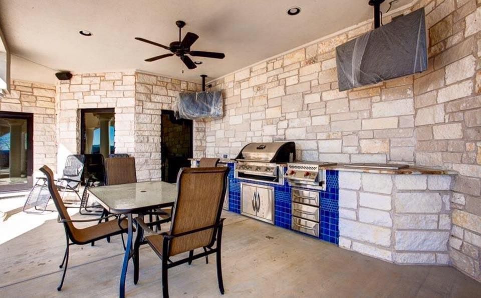 Outdoor patio with stone walls, table and chairs, ceiling fan, and built-in blue grill