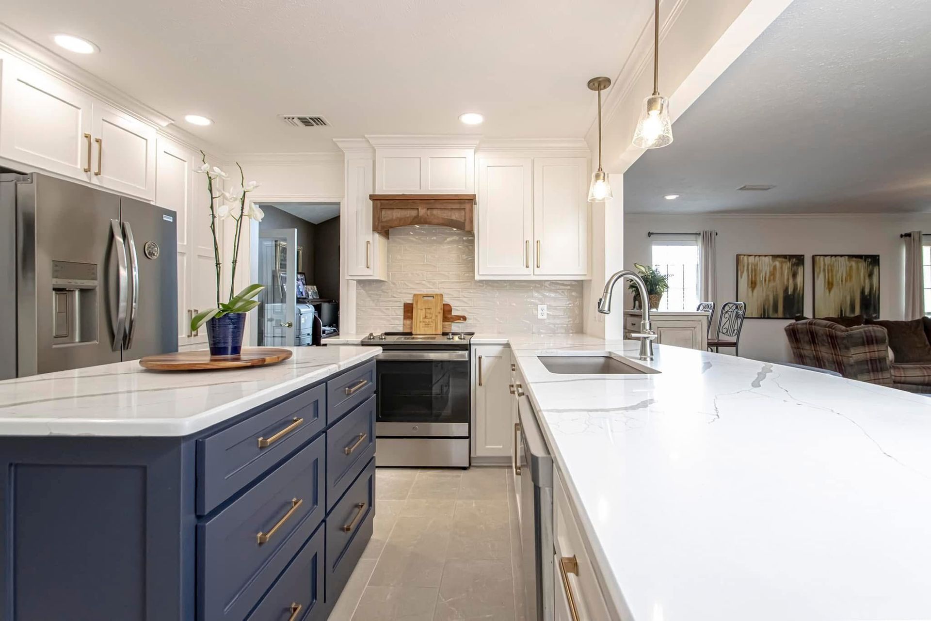 Bright modern kitchen with white countertops, blue cabinets, stainless steel fridge, and open living room beyond