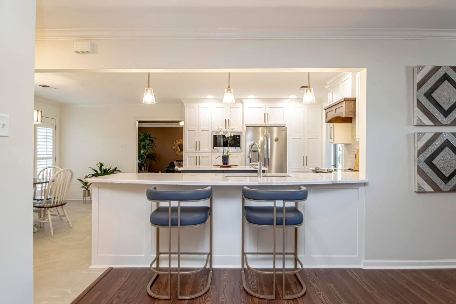 Bright kitchen pass-through with white cabinets, island, and two blue bar stools over dark wood flooring