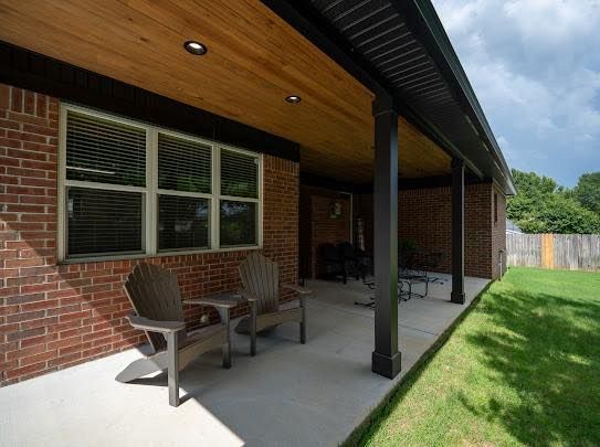 Covered brick patio with chairs, wood ceiling, and lawn beside a house