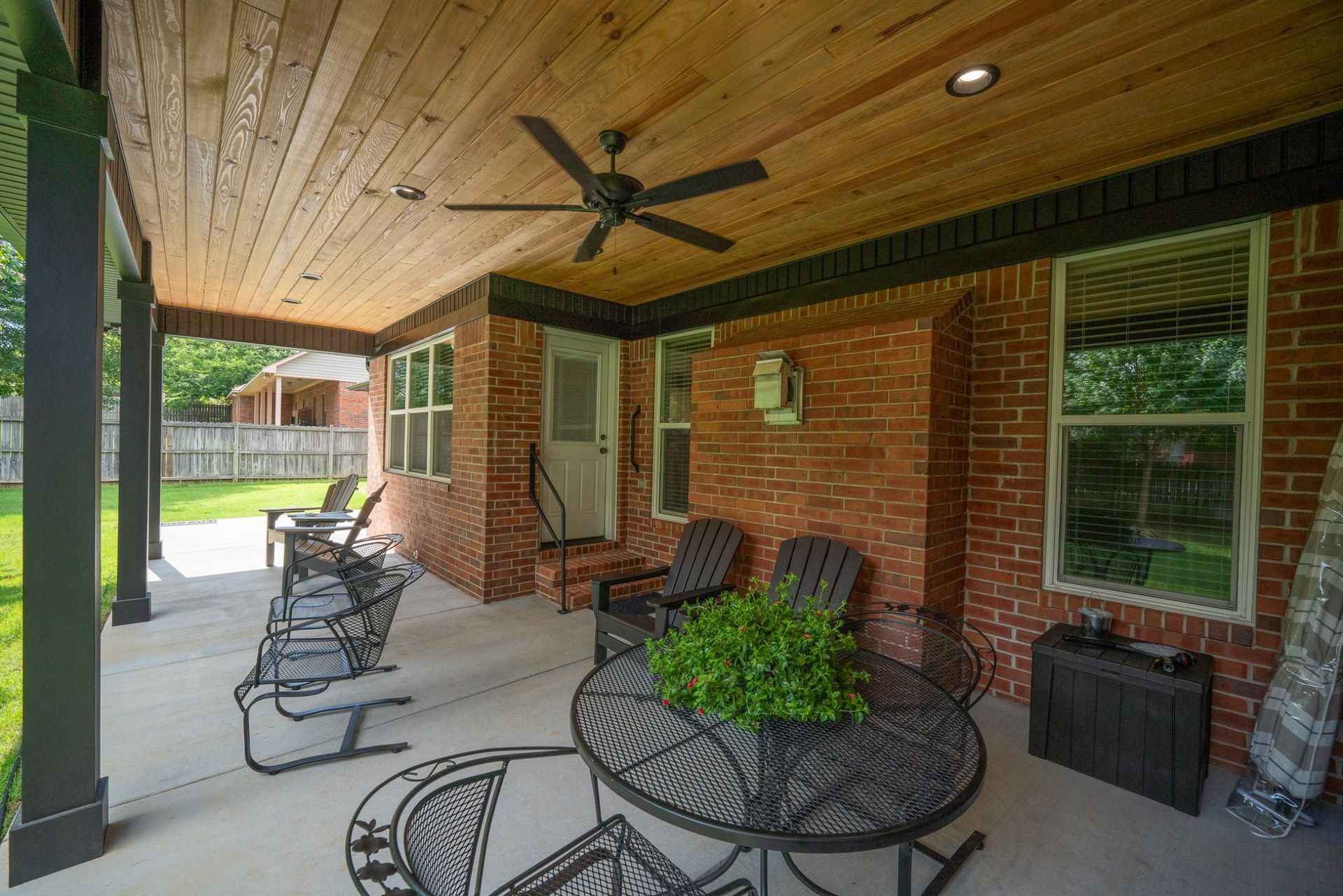 Covered brick patio with ceiling fan, metal chairs, and table beside a grassy yard