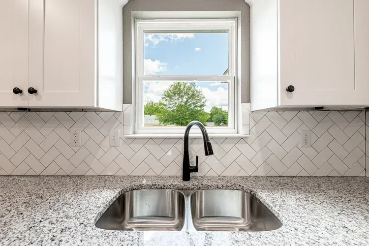 Bright kitchen sink with black faucet, speckled countertop, white cabinets, and a window above the sink.