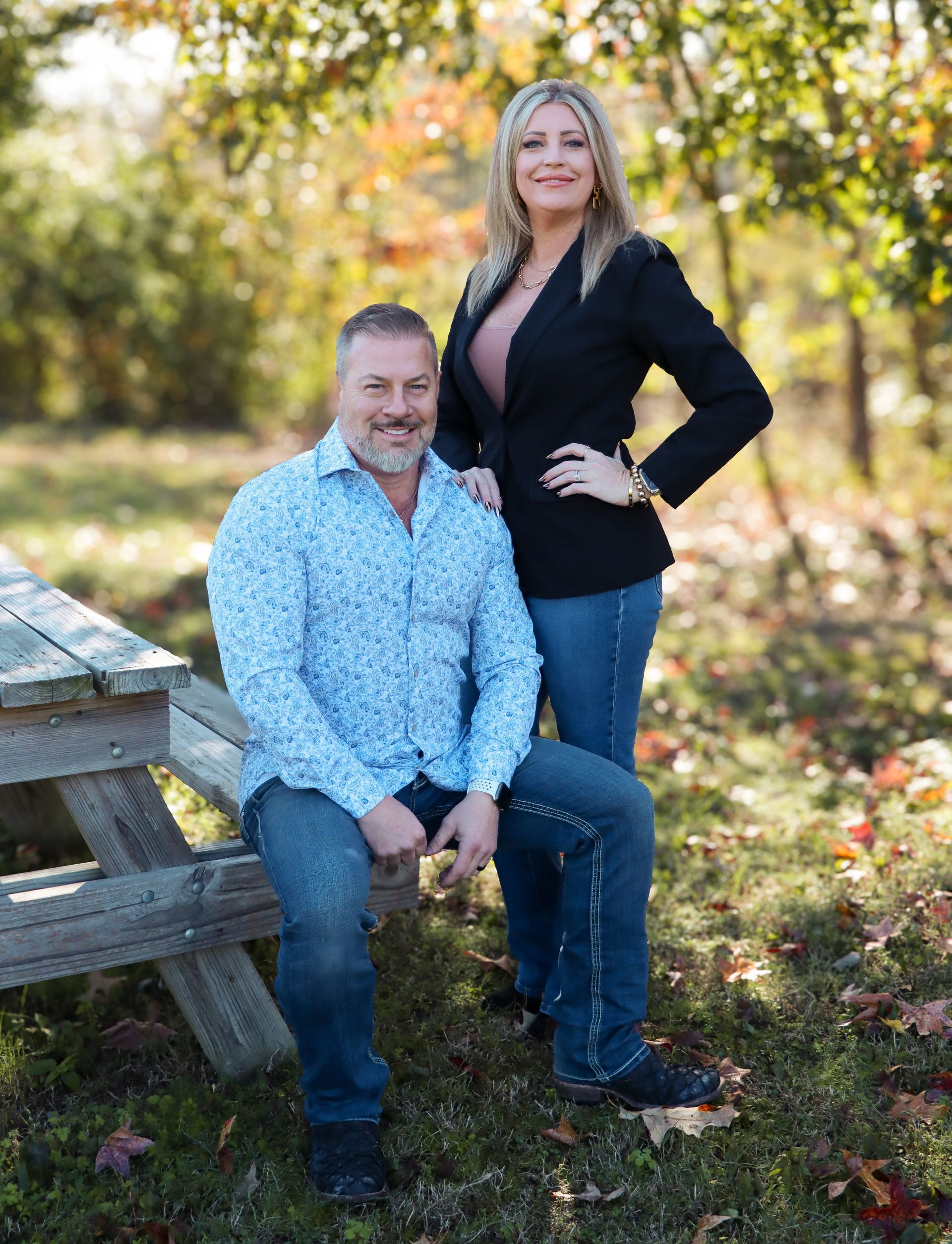 Two people pose outdoors by a wooden bench amid autumn foliage, smiling at the camera.