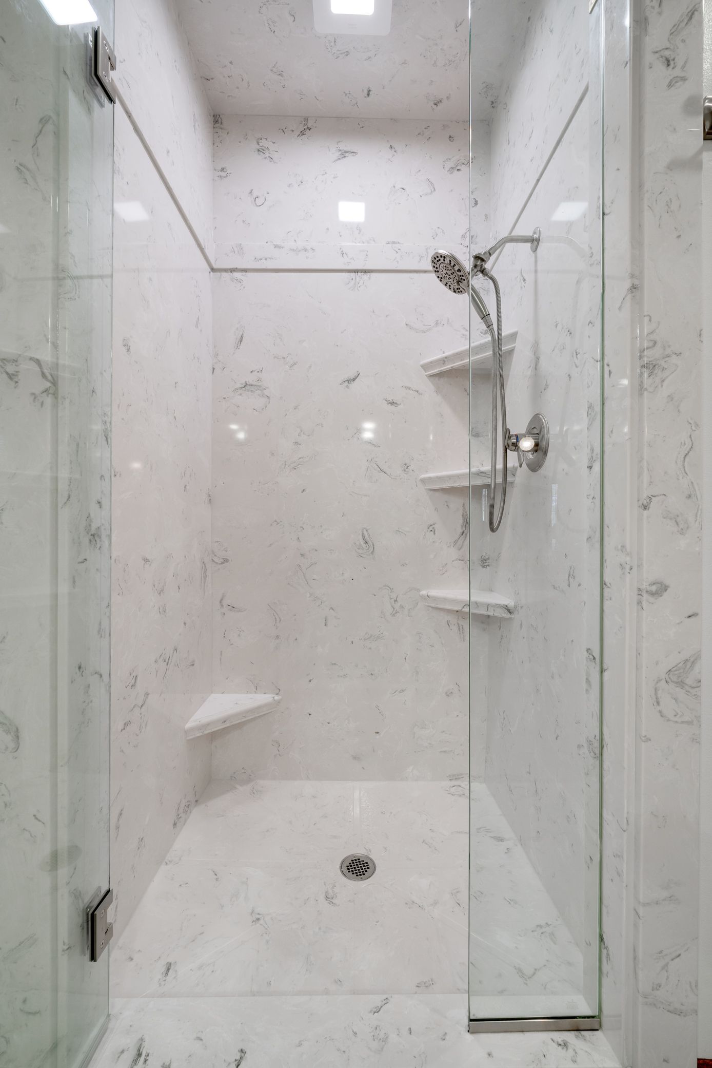 Compact white tiled shower stall with glass door, corner shelves, and handheld showerhead.
