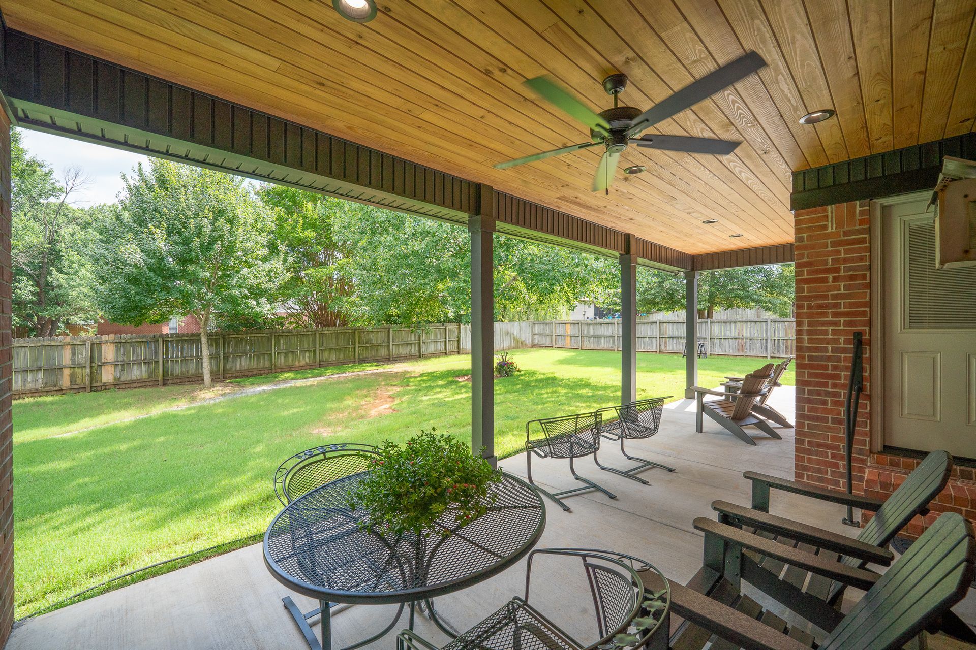 Covered patio with ceiling fan, outdoor chairs, and round table overlooking a green backyard.