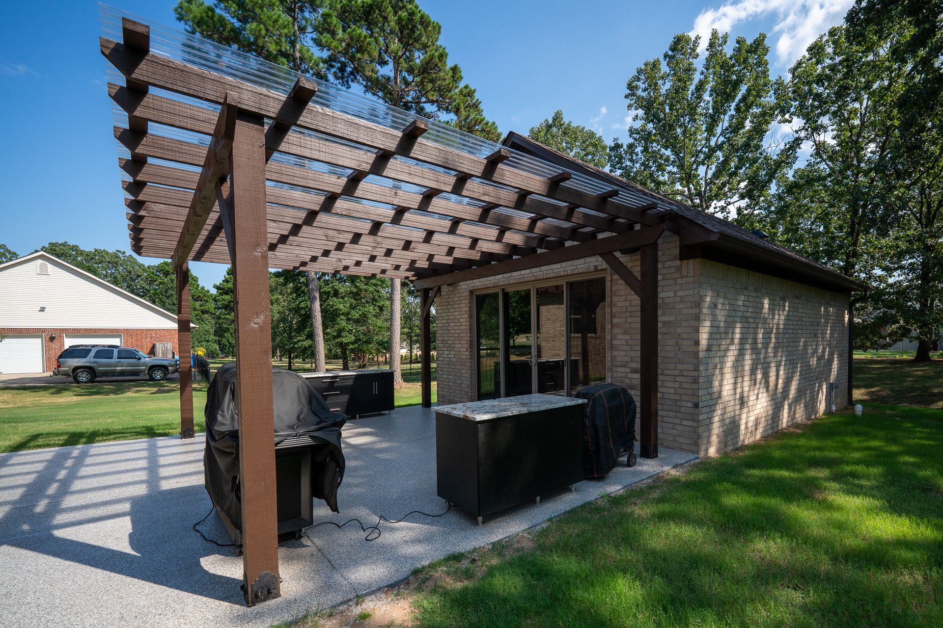 Covered backyard patio with wooden pergola, grill, and outdoor dining area beside a brick house